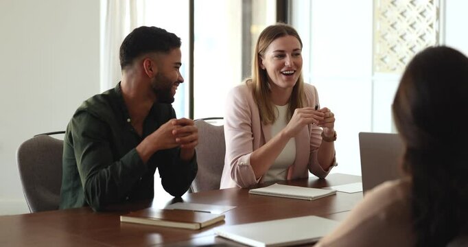 Two positive diverse young company colleagues discussing work strategy at team meeting, speaking, smiling, laughing, nodding, sitting at large table, brainstorming on ideas with business group