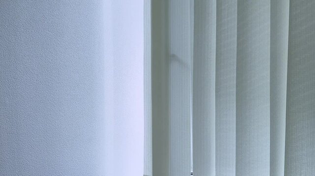 Vertical white blinds swaying in wind or draft near open window. Light and shadows on wall. Light and shadows on wall