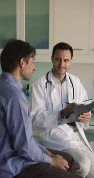 Mature male doctor in white uniform and stethoscope on neck talk to man patient, discuss concerns, writes health complaints, filling medical form card. General check-up, healthcare, medical insurance