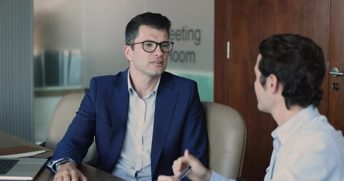 Serious middle aged company executive meeting with younger business consultant, consulting expert, advisor, nodding. Business leader, director listening to employee at workplace table