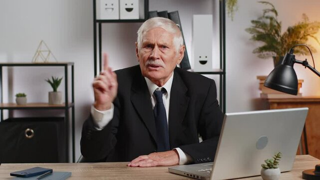 Caucasian senior businessman with laptop shakes finger and saying No be careful scolding and giving advice to avoid mistake disapproval sign at home office. Freelancer grandfather sitting at desk.