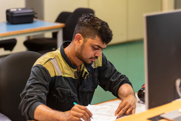 The marine engineer officer sits in the control room of a merchant vessel, focused on recording key operational parameters in the engine logbook, surrounded by monitors displaying system data.