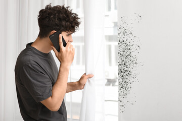 Young man talking by mobile phone with mold on light wall near window at home