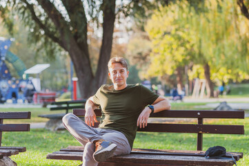 A cheerful young man is sitting alone in the park on the bench and enjoying the autumn sun