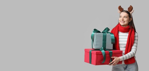 Beautiful young woman in reindeer horns and with gift boxes on grey background with space for text