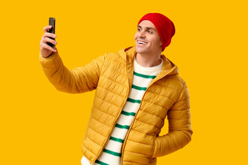 Young man in stylish puffer jacket and hat taking selfie on yellow background