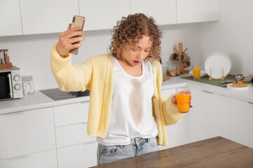 Shocked young woman with mobile phone spilled juice on her clothes in kitchen