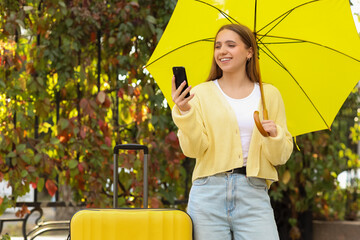 Female tourist with umbrella and suitcase using mobile phone in park on autumn day