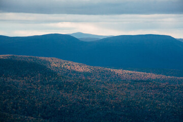 Vast mountain landscape under dramatic clouds at sunset, showcasing layered hills and illuminated autumn foliage in a remote wilderness setting