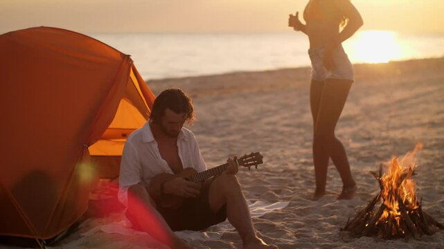 A serene beach scene at sunset with a man playing the ukulele and a woman dancing near a campfire. Captures the essence of carefree summer evenings, perfect for travel and lifestyle concepts.