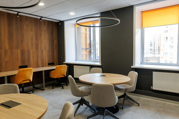 Empty working and meeting area in an open office. White round wooden table surrounded by chairs in grey and yellow colors. Big window background