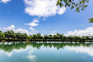 White clouds float in the sky, reflecting beautifully on the water's surface. Trees line the shore, creating a peaceful atmosphere. This photo was taken in Karen State, Myanmar in August.