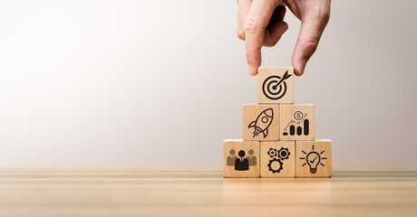 Hand arranging wooden blocks with business icons symbolizing startup growth, teamwork, financial success, goal achievement, representing the steps to build a successful, innovative business strategy