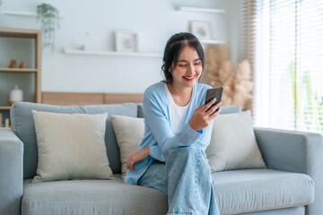 Smiling pretty young Asian woman sitting on cozy couch, using funny mobile apps in living room. Woman at home, Doing online shopping, Messaging friends, Posting on social media