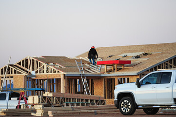 A construction worker atop the structure of a wooden home under construction, wearing a tool belt...