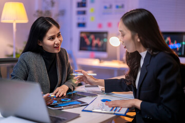 Two women are sitting at a desk with laptops and papers