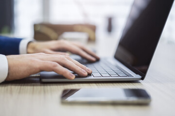 With an office scene appearing softly blurred in the backdrop, the picture shows a close-up of male hands typing on a contemporary laptop keyboard