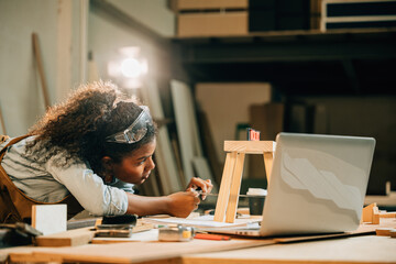 African American carpenter woman works on a wooden project using a laptop. utilize modern...