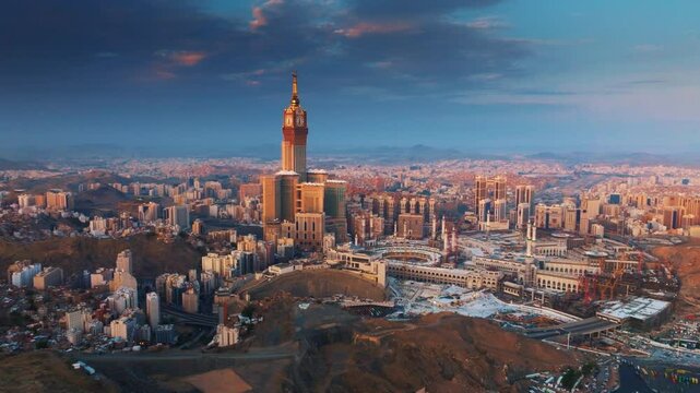 Aerial view of the Abraj Al-Bait Clock Tower and Grand Mosque in Mecca illuminated by the golden light of sunrise, surrounded by the sprawling holy city and mountains