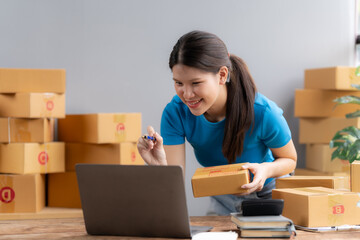 Online Business Owner: A young woman, full of energy and enthusiasm, manages her online business from home. She holds a package while checking her laptop, surrounded by stacks of boxes.