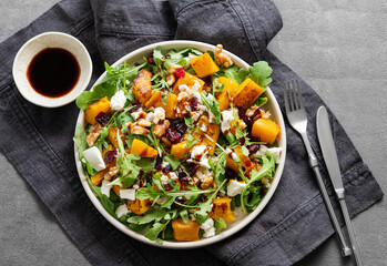 Autumn salad with pumpkin, arugula, feta cheese, nuts and dried berries in a plate on a napkin on dark background