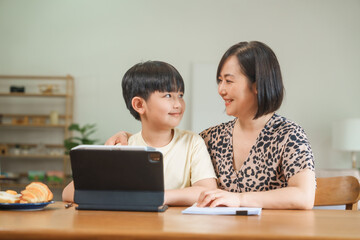 A mother teaches her son to study online at home, using a digital tablet for learning. They sit together at a table with snacks, promoting homeschooling and family bonding.