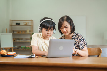 A mother teaches her son to study online at home, using a digital tablet for learning. They sit together at a table with snacks, promoting homeschooling and family bonding.