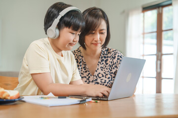 A mother teaches her son to study online at home, using a digital tablet for learning. They sit together at a table with snacks, promoting homeschooling and family bonding.