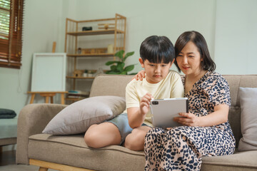 An Asian mother and son sit at the table and sofa, sharing time together while looking at a computer, tablet, and phone. They bond through technology and family activities