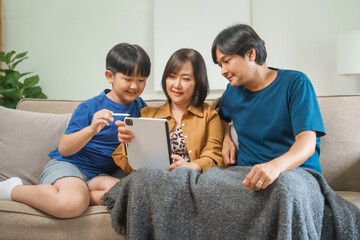 Asian parents and children sit together on a sofa at home, laughing and enjoying family time while...