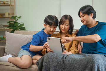 Fototapeta premium Asian parents and children sit together on a sofa at home, laughing and enjoying family time while using various devices like laptops, phones, and tablets, showcasing a tech-savvy lifestyle
