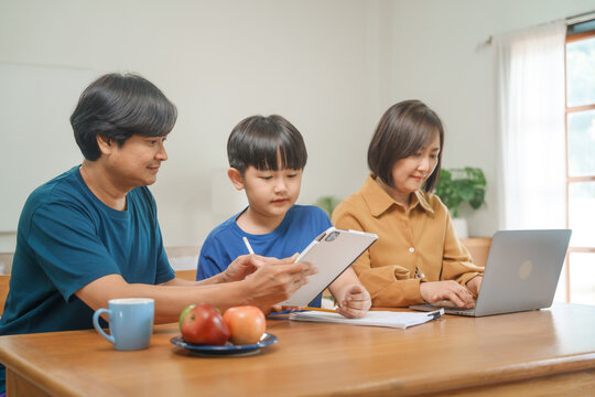 Asian parents and their children sit together at the table in the morning, studying online using tablets and computers, fostering a productive and focused family learning environment