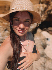 A cheerful young woman in a straw hat and bikini smiles while taking a selfie at the beach. Surrounded by the coastline and natural beauty, she embodies summer relaxation and enjoyment in Melbourne