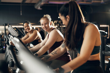 Fit group of young diverse women in sportswear smiling while riding on a stationary bikes during a cardio workout session together in a health club