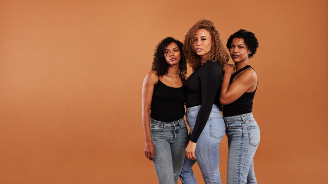 Diverse women in jeans and black tops standing on an orange background. They all have curly hair of different lengths and look focused directly into the camera
