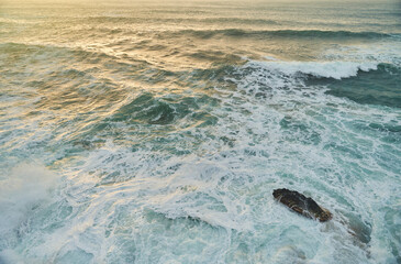 A closeup shot of the ocean with wind waves crashing onto the shore, creating a beautiful pattern of furrows in the water. The landscape is enhanced by cumulus clouds in the sky