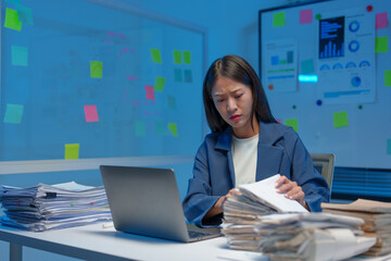 A tired, sleepy, bored businesswoman sits at a desk for a long time, working with a pile of papers. Lots of documents piled up on desks and laptop computers. Office syndrome concept.