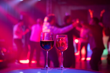Two elegant wine glasses on a table with people dancing in background under colorful party lights, creating a vibrant and lively atmosphere.