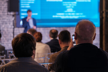 Audience members attentively watch a speaker during conference, capturing moments on their phones in a dimly lit venue.