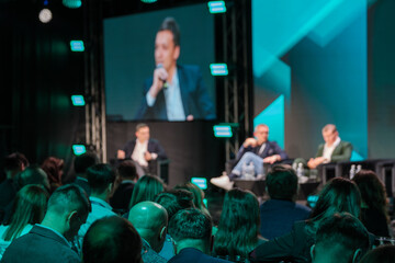 Audience attentively listening to panel discussion at business conference. Blurred stage and vibrant lights create engaging atmosphere for networking and learning.