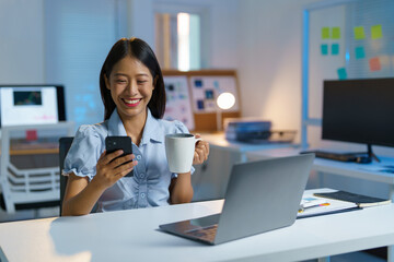 Asian woman, businesswoman sitting texting FaceTime or watching video on her smartphone while holding a cup of coffee drinking relaxing while working at home office. Work lifestyle concept.