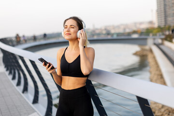 A young woman enjoys a peaceful moment listening to music on a tranquil bridge during sunset by the water