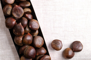 Fototapeta premium Sweet chestnuts in cardboard punnet on linen, from above, with empty space for text. Castanea sativa, raw, whole and unshelled. Can be eaten raw, candied, cooked, or roasted as warm snack on streets.