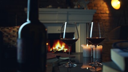 Glass of red wine, book and candle on table in dark room with fireplace. Cozy warm home in winter.
