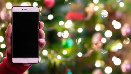 Woman holding mobile phone and showing screen with christmas tree lights on the background. Concept of online service in holidays.