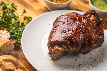 Baked pork knuckle on a white plate on a wooden background or table with bread, fresh cucumbers, parsley and mustard. View from above.