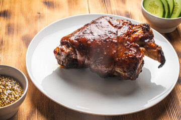 Baked pork knuckle on a white plate on a wooden background or table with bread, fresh cucumbers, parsley and mustard. View from above.