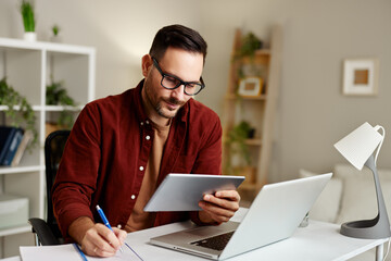 Businessman taking notes while working remotely from home office using laptop and digital tablet