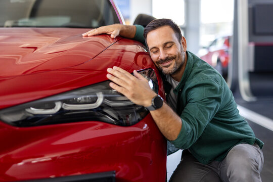 Happy young man hugging his new car in showroom. Satisfied guy with closed eyes embracing the hood of the automobile. Dreaming man lying on car bonnet hugging it.
