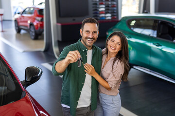 Visiting car dealership. Beautiful couple is holding a key of their new car, looking at camera and...